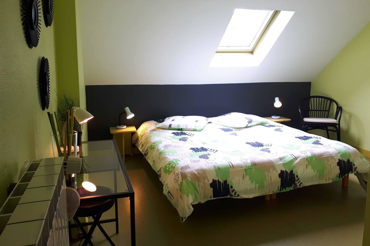 A bedroom with a skylight, a bed with a floral bedspread, a desk with a keyboard, a chair, and a nightstand with lamps.