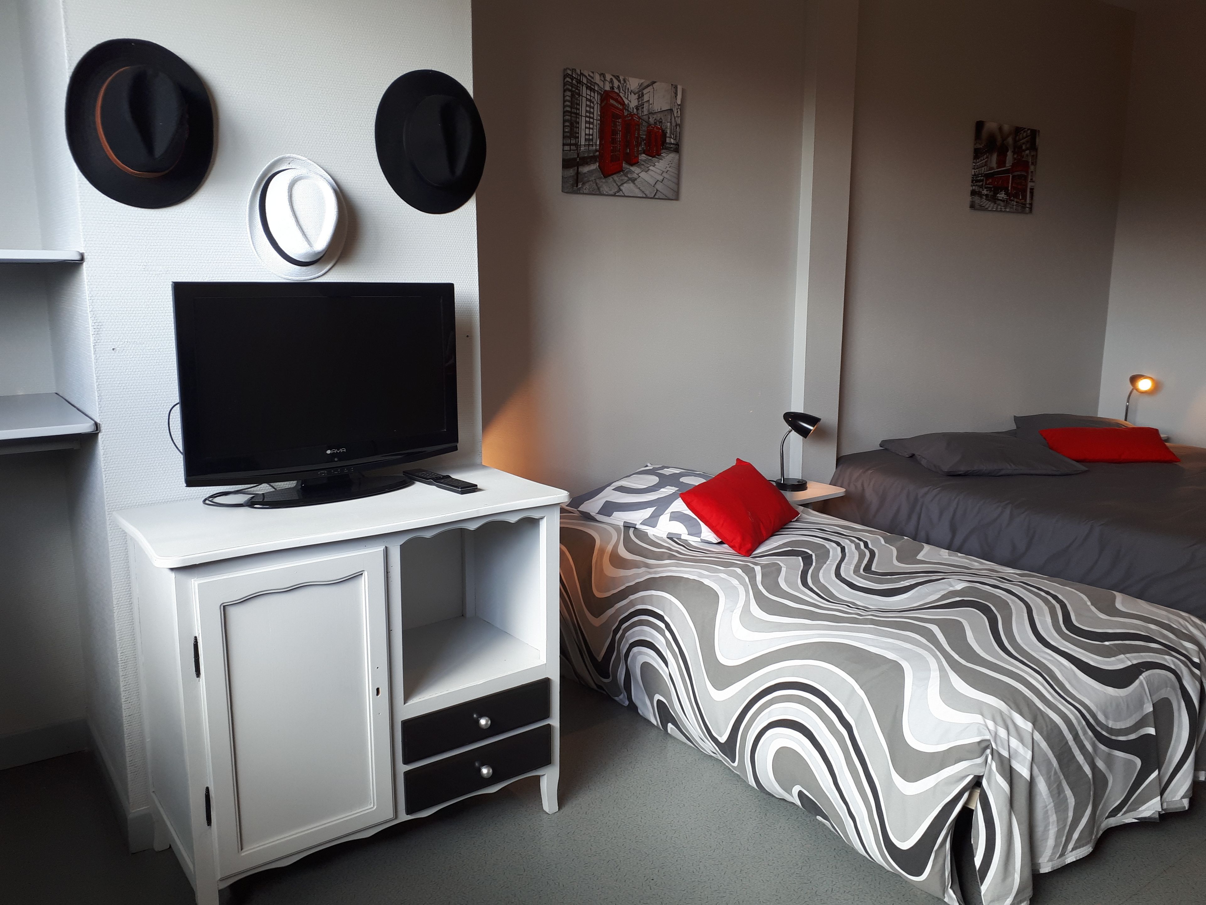 Un dormitorio con una televisión sobre un escritorio blanco, dos camas con ropa de cama estampada y sombreros colgados en la pared.