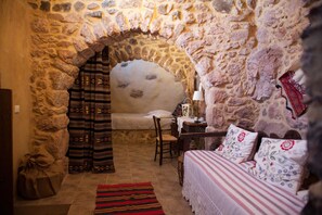 A stone-walled room with a bed, a chair, a table with a lamp, and a patterned rug. - Sitanos Forgotten Neighborhood (Sitia)
