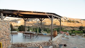 Une terrasse extérieure dotée d’une pergola en bois, de murs en pierre, d’une table avec des chaises et offrant une vue sur un paysage montagneux.