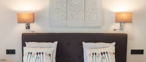 Una habitación de hotel con cabecera oscura, dos lámparas de noche, un adorno de pared y dos almohadas con estampado floral.