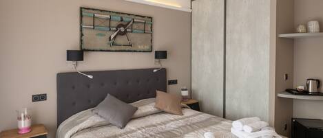 A modern hotel room with a large bed, a grey headboard, a painting above the bed, a bedside table with a lamp, and a wardrobe.
