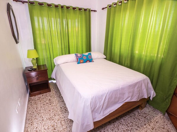 A bedroom with a bed, green curtains, a nightstand with a lamp, and a carpeted floor.