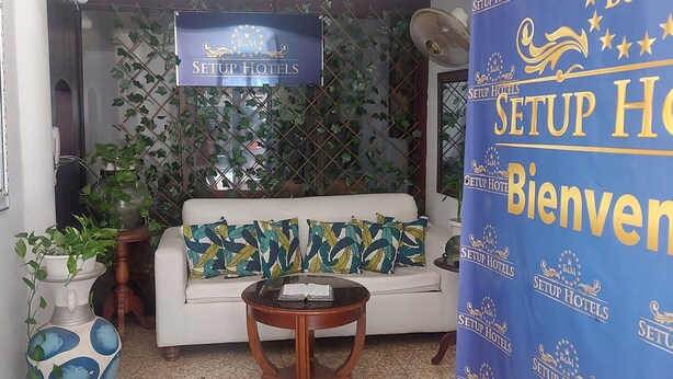 Um hall de hotel com um sofá branco, almofadas estampadas, uma mesinha de madeira e plantas em vasos.