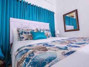 A neatly made bed with a patterned bedspread, a white headboard, and a blue curtain. - Setup Hotels (Santo Domingo Este)
