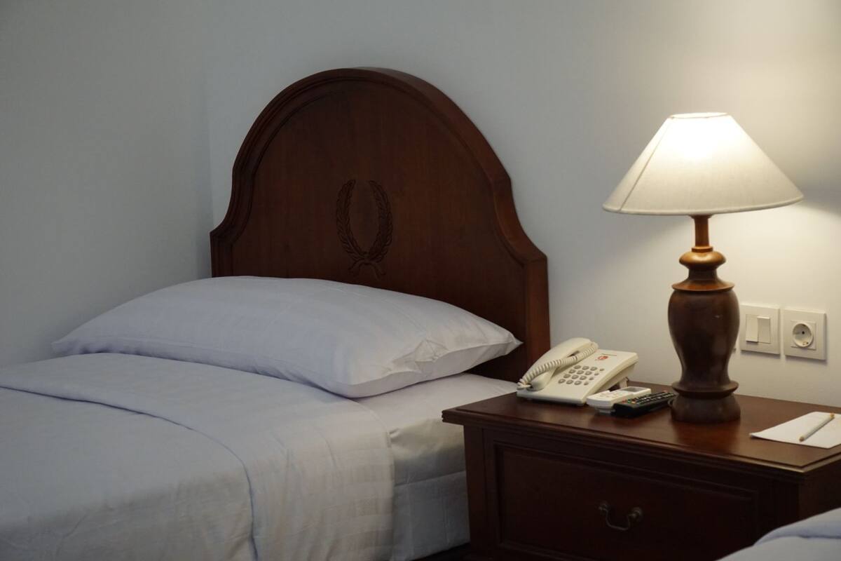 Uma cama bem arrumada com lençóis brancos, uma cabeceira de madeira com desenho de grinalda de loureiro, um abajur, um telefone e uma mesa de cabeceira com uma gaveta.