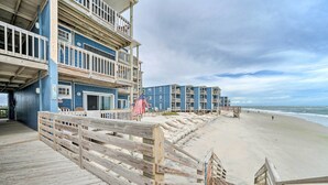 Ein hölzerner Steg führt zu einem Strand, gesäumt von einer Reihe blauer Gebäude mit Balkonen.