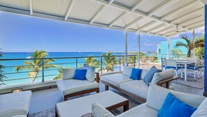 A covered outdoor seating area with white sofas and blue cushions, overlooking a beach with palm trees and a clear blue sky.