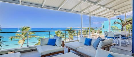 A covered outdoor seating area with white sofas and blue cushions, overlooking a beach with palm trees and a clear blue sky.