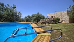 A swimming pool with a wooden deck and metal ladder, surrounded by greenery and a stone building.