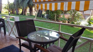 A patio with a round glass table and two chairs, a striped awning, and a view of a well-maintained garden with palm trees. - CALA DEL PI RESIDENTIAL APARTMENT (TREUMAL CENTER) PLATJA D¨ARO (Platja D Aro)