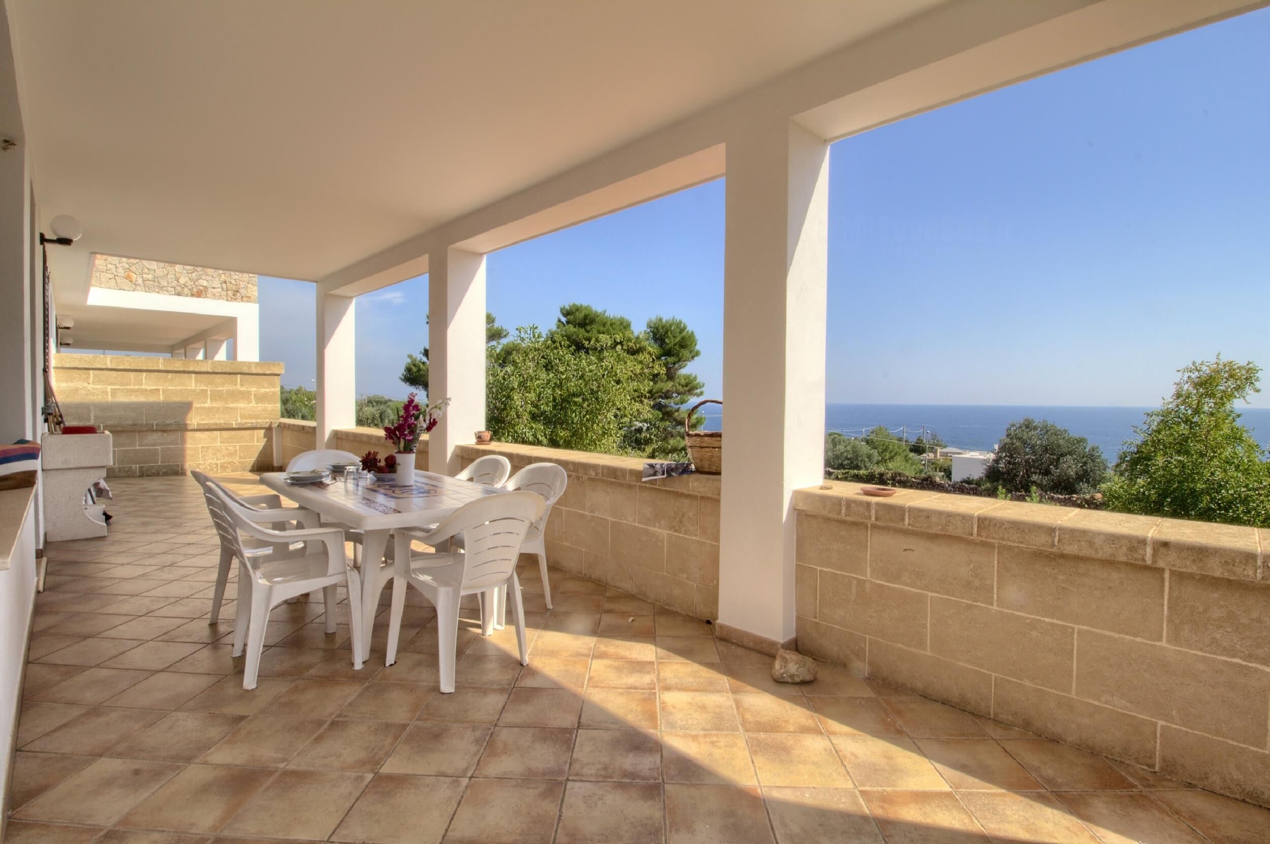 A covered patio with a table and chairs, a stone wall, and a view of the sea.