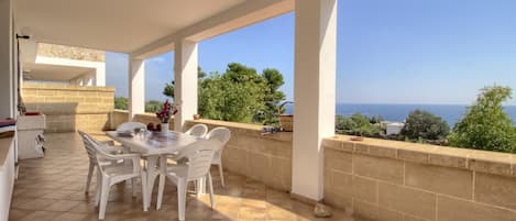 A covered patio with a table and chairs, a stone wall, and a view of the sea.