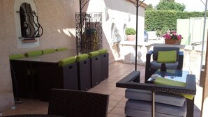 Een terras buiten met rieten meubilair, een ronde tafel en een zithoek met kussens.