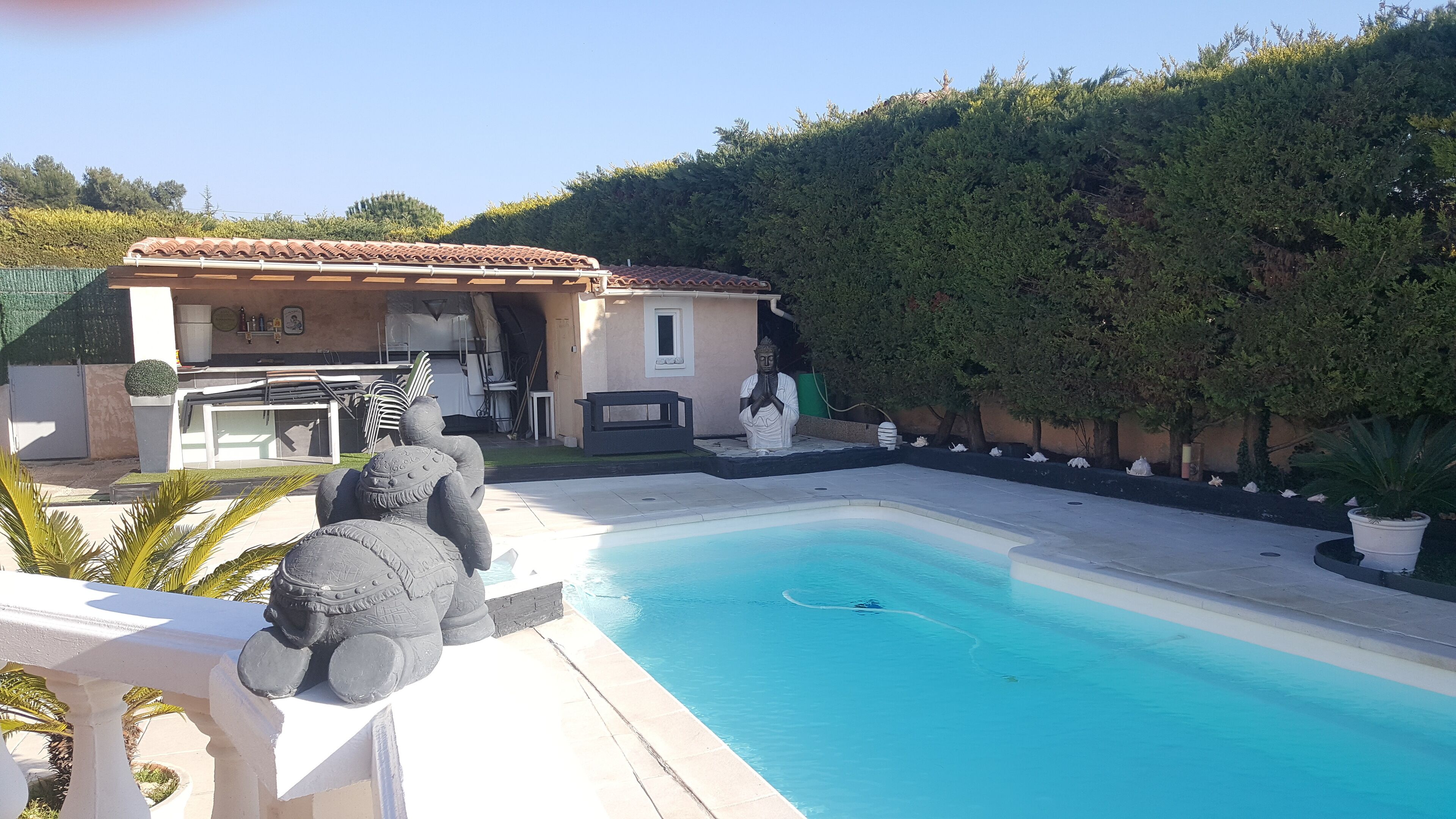 A swimming pool with a statue in the foreground, a building with a tiled roof, and a hedge in the background.