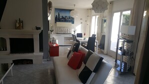 Een moderne woonkamer met een open haard, een bank met rode kussens, een eethoek met stoelen en een groot raam met gordijnen.