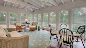 A sunlit room with wicker furniture, a ceiling fan, and large windows offering a view of greenery outside.