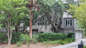 A two-story house with a wooden staircase leading to the upper level, surrounded by lush greenery and a mailbox on the sidewalk.