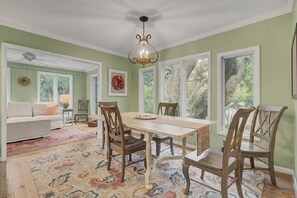 Un comedor con mesa y sillas de madera, una lámpara de araña y ventanas amplias con vista a árboles.