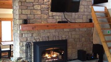 A stone fireplace with a TV mounted above it, a wooden staircase to the right, and a sofa in the foreground.