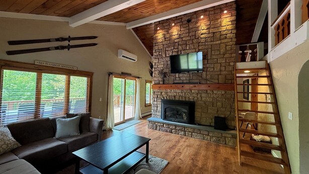 A cozy living room with a stone fireplace, a flat-screen TV mounted on the wall, a wooden ceiling, and a staircase leading to an upper level. - Beautiful Chalet Style Home with Seasonal Mountains Views in North Conway, NH (North Conway)