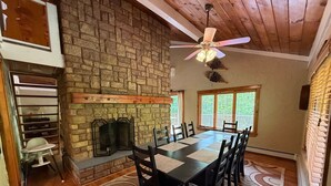 A dining area with a stone fireplace, a ceiling fan, and a large window with blinds. - Beautiful Chalet Style Home with Seasonal Mountains Views in North Conway, NH (North Conway)