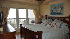 A bedroom with a wooden bed, a nightstand with a lamp, a painting of boats on the wall, and a view of the outdoors through large windows. - Beachfront condo: Create some memories! (South Padre Island)