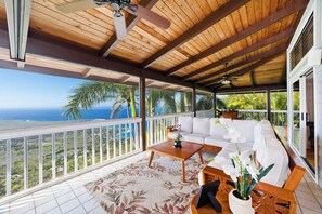 A covered balcony with a wooden ceiling, white cushioned seating, a wooden table, and a view of the ocean. - Maka Nui Estate/ Luxury Big Island Ocean View Home Above Kealakekua Bay (Captain Cook)
