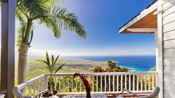 A wooden deck with white railings, two wicker chairs, a potted plant, and a view of the ocean and landscape. - Maka Nui Estate/ Luxury Big Island Ocean View Home Above Kealakekua Bay (Captain Cook)