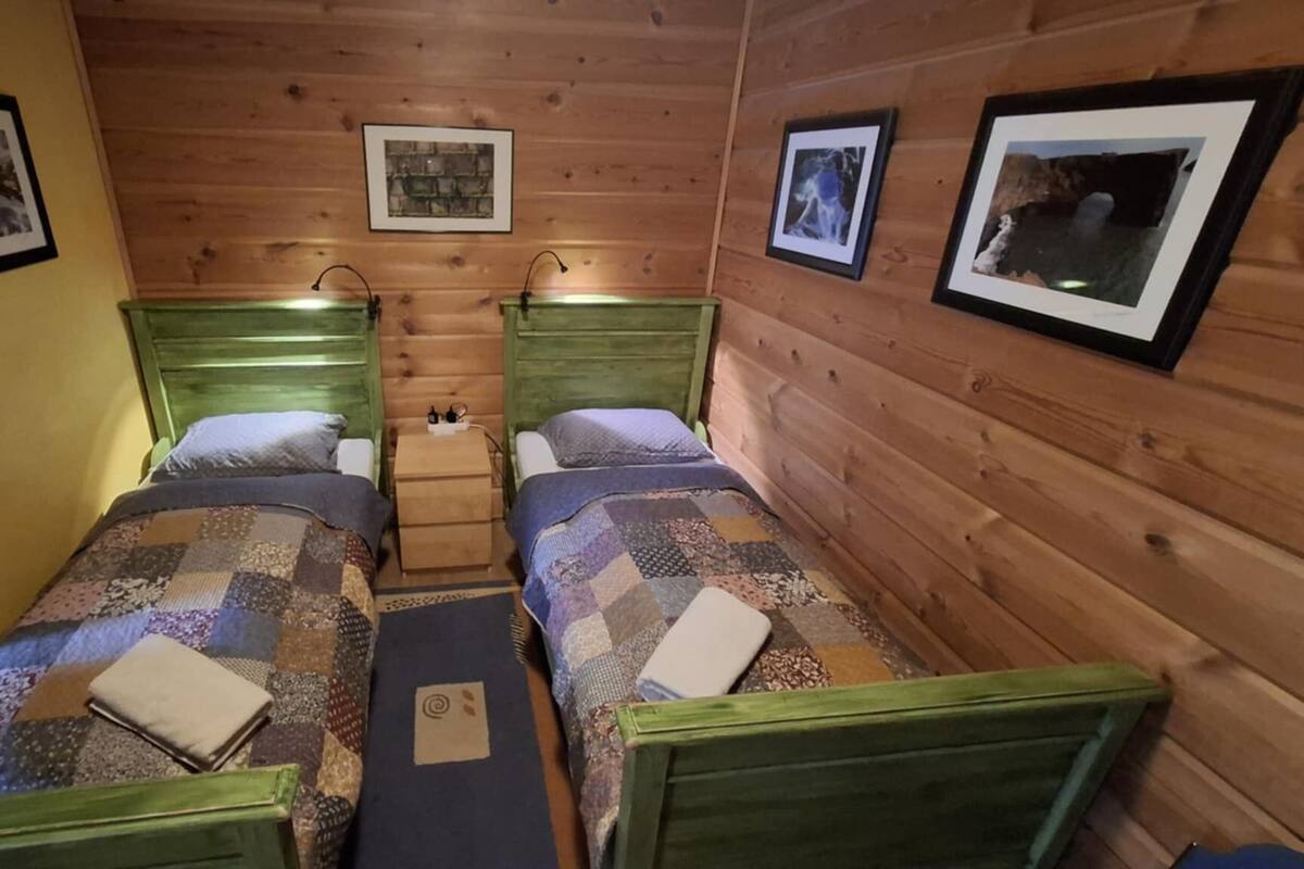 Una habitación con dos camas de madera, cada una con cabecero y almohada. Hay cuadros enmarcados en las paredes y una mesita entre las camas.