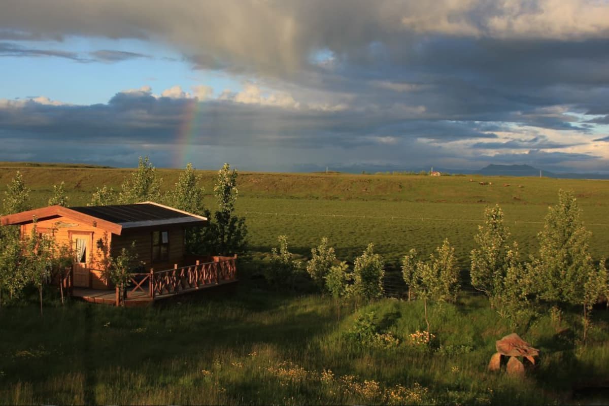 Una cabaña rústica con terraza de madera, rodeada por un campo cubierto de hierba y árboles, bajo un cielo nublado.