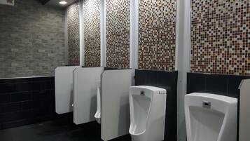 A public restroom with multiple urinals, tiled walls, and a control panel on the wall.