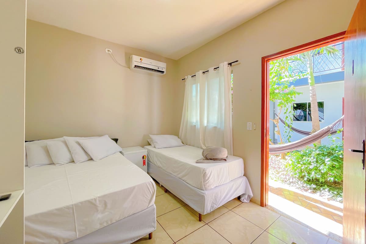 A hotel room with two beds, an air conditioning unit, a window with sheer curtains, and a view of a building and greenery outside.
