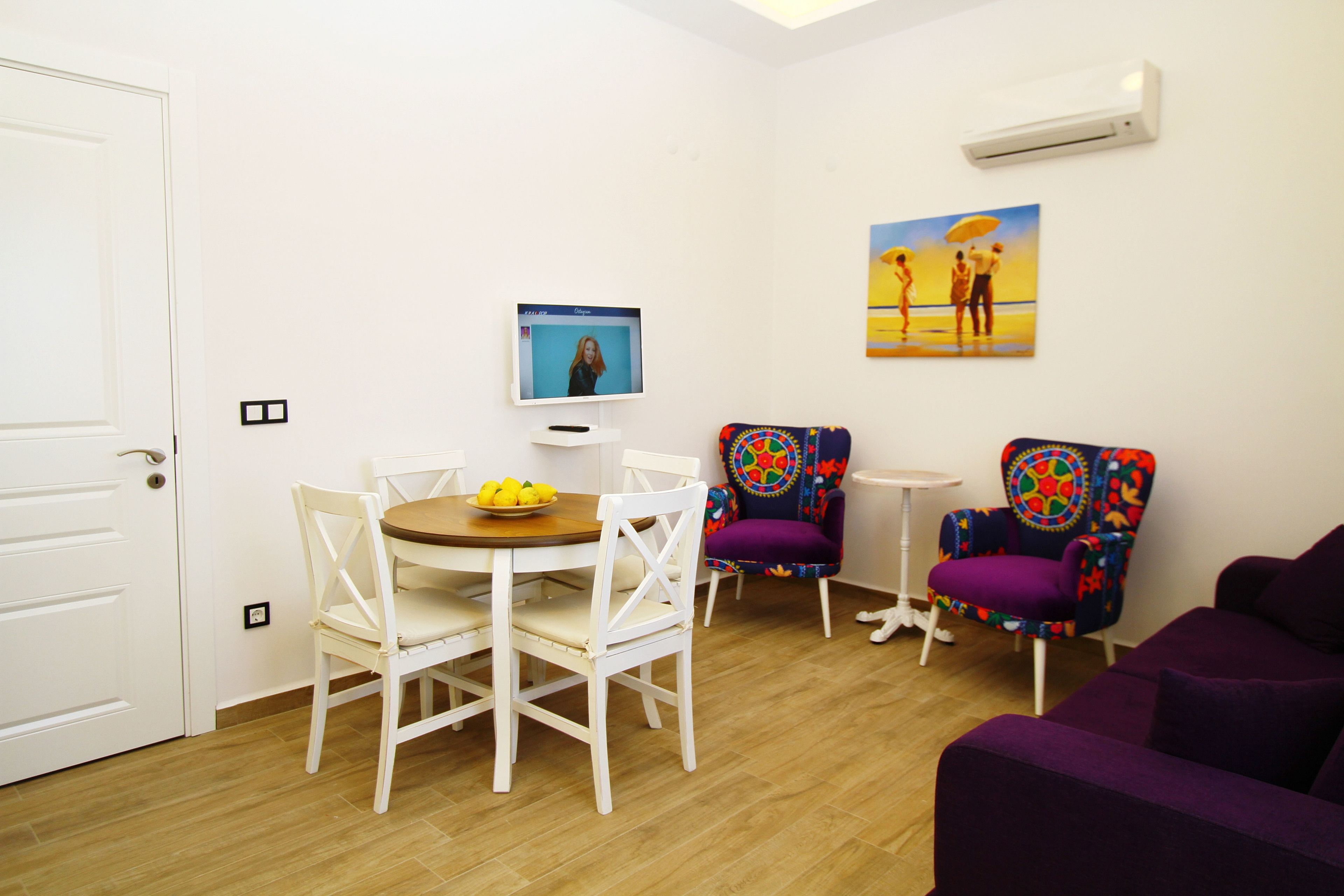 A room with a dining table, chairs, a purple sofa, a colorful armchair, a small round table, and a painting on the wall.