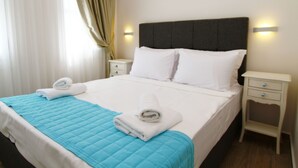 A neatly made bed with white linens and a blue quilt, two white pillows, and two rolled towels on the bed. There is a small bedside table with a drawer and a wall-mounted light. - Cesme Kale Tas Apart (Cesme)