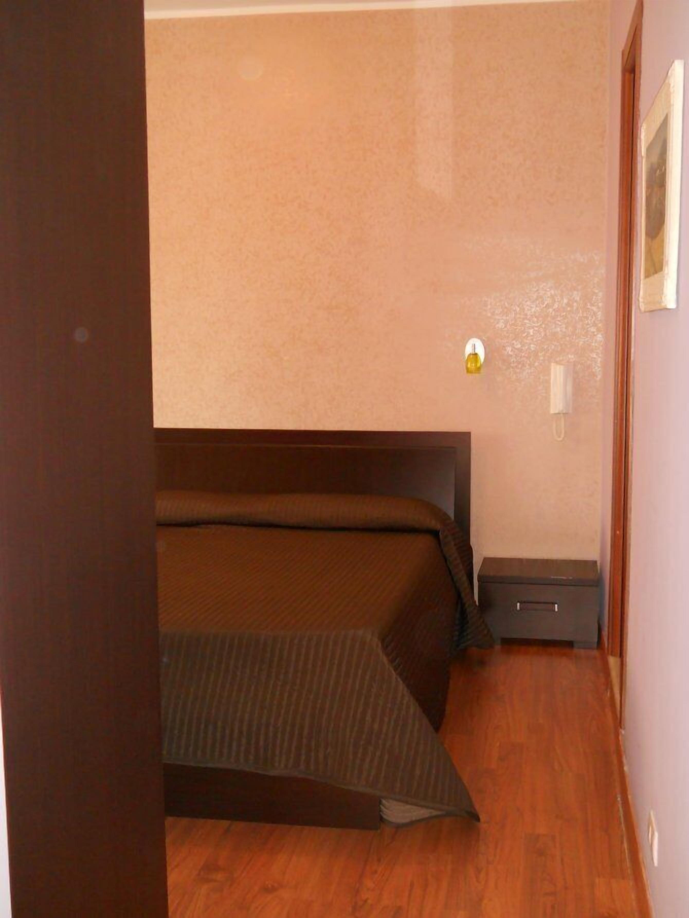 A small, compact room with a single bed, a nightstand, and a wall-mounted light fixture.