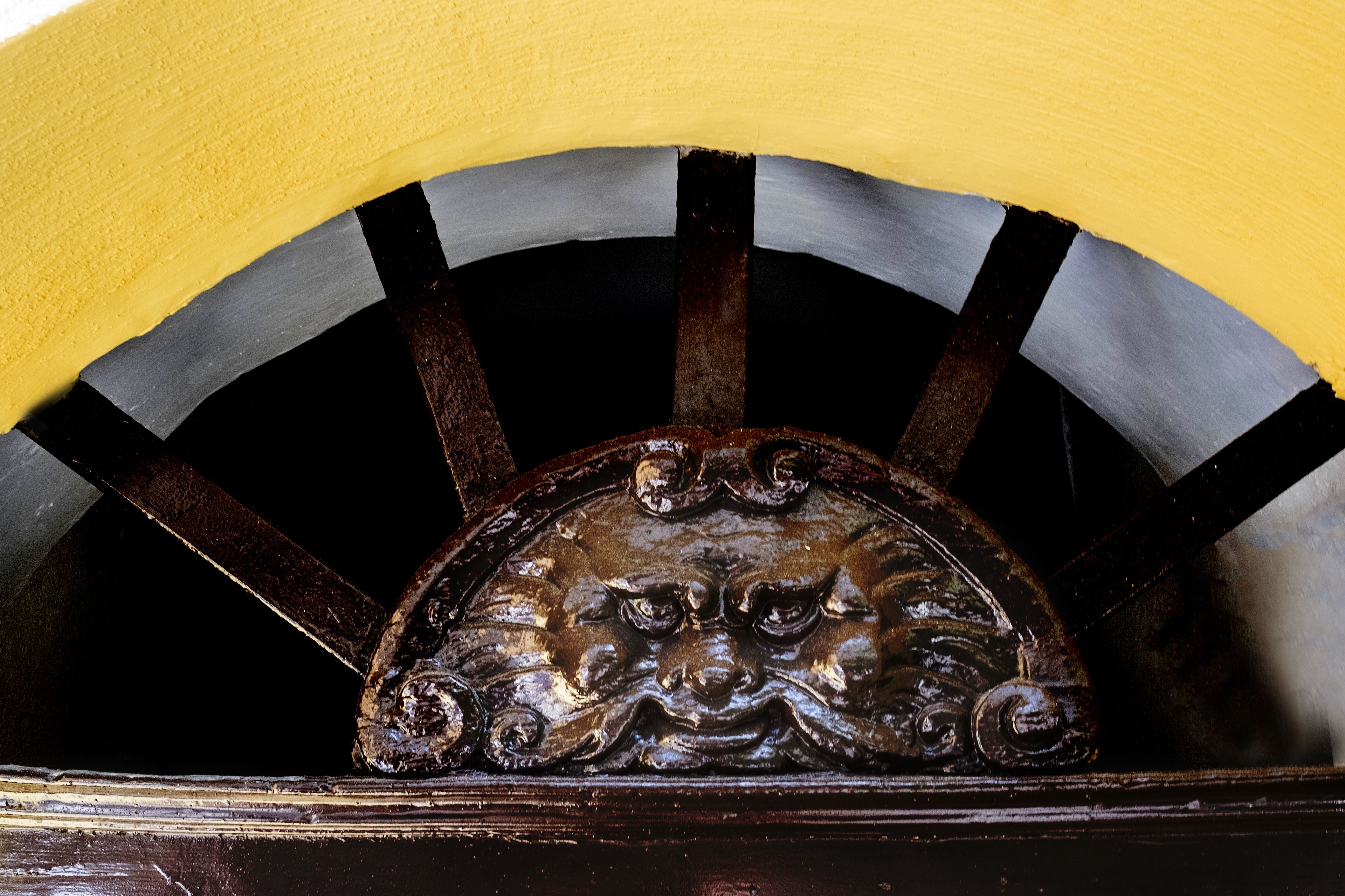 Detalle de una puerta con un escudo metálico decorativo y un arco a rayas amarillas y negras.