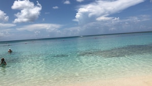 Uma praia com águas turquesa cristalinas, areal arenoso e algumas pessoas a nadar.