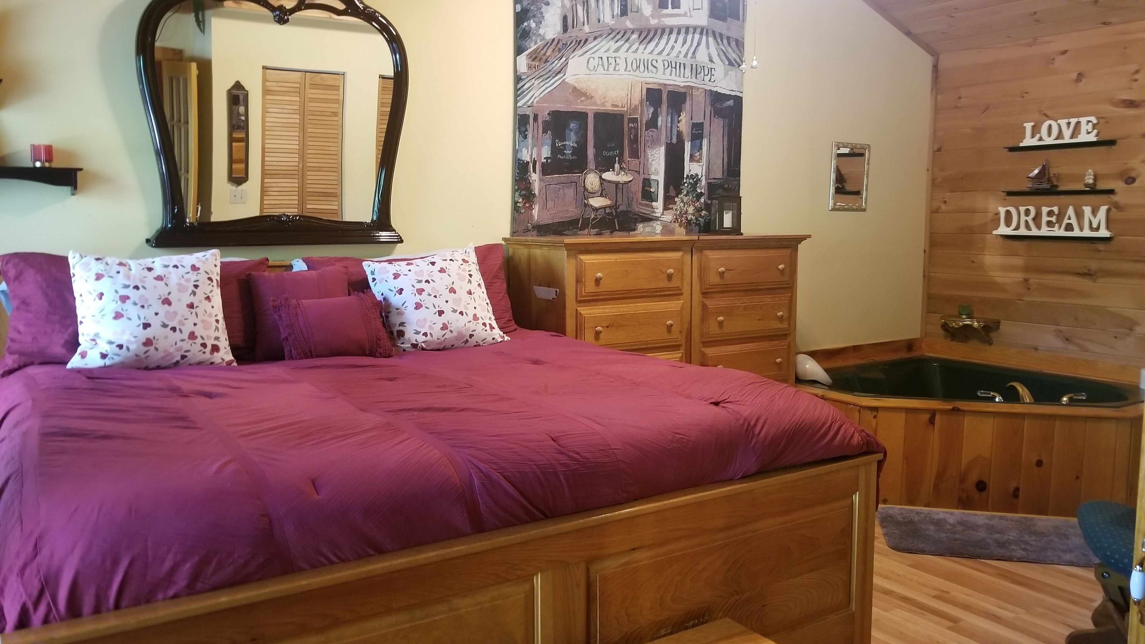 A bedroom with a wooden bed, a large mirror, a dresser, a wall-mounted shelf with decorative items, and a poster of a café.