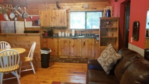 A cozy cabin interior with a wooden dining area, a kitchen with wooden cabinets, and a leather sofa with a patterned cushion.