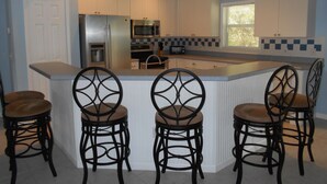 Una cocina con una isla central, cuatro taburetes de bar, electrodomésticos de acero inoxidable y un ventanal con persianas.