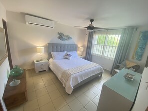 A bedroom with a bed, bedside tables, an air conditioner, a ceiling fan, and a window with curtains. - Beachfront Paradise With Amazing Ocean Views! Private access to Beach. Wifi (Manati)