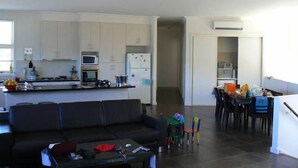 A modern kitchen with white cabinets, a microwave, and a refrigerator. There is a dining area with a table and chairs, and a living room with a black sofa and a coffee table.
