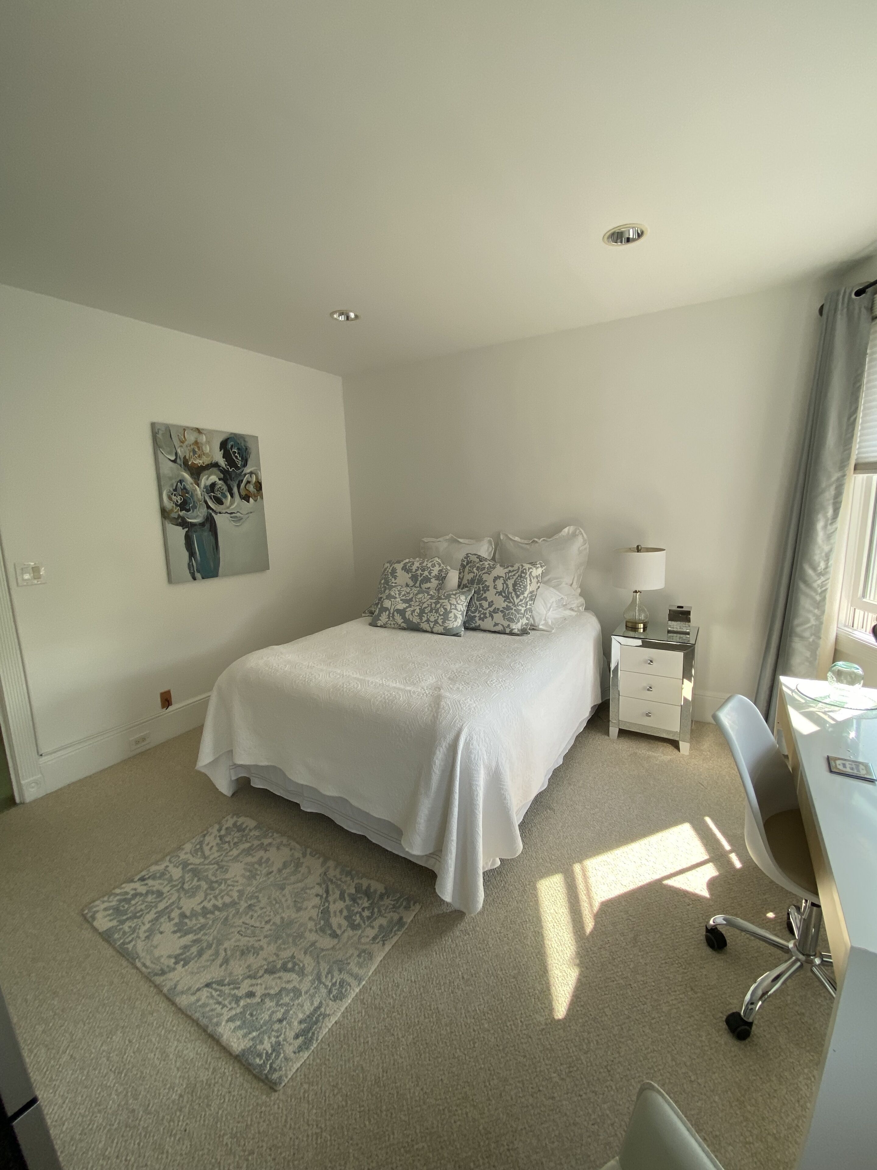 A bedroom with a bed, bedside tables, a desk, a chair, a rug, a painting, and a window with curtains.