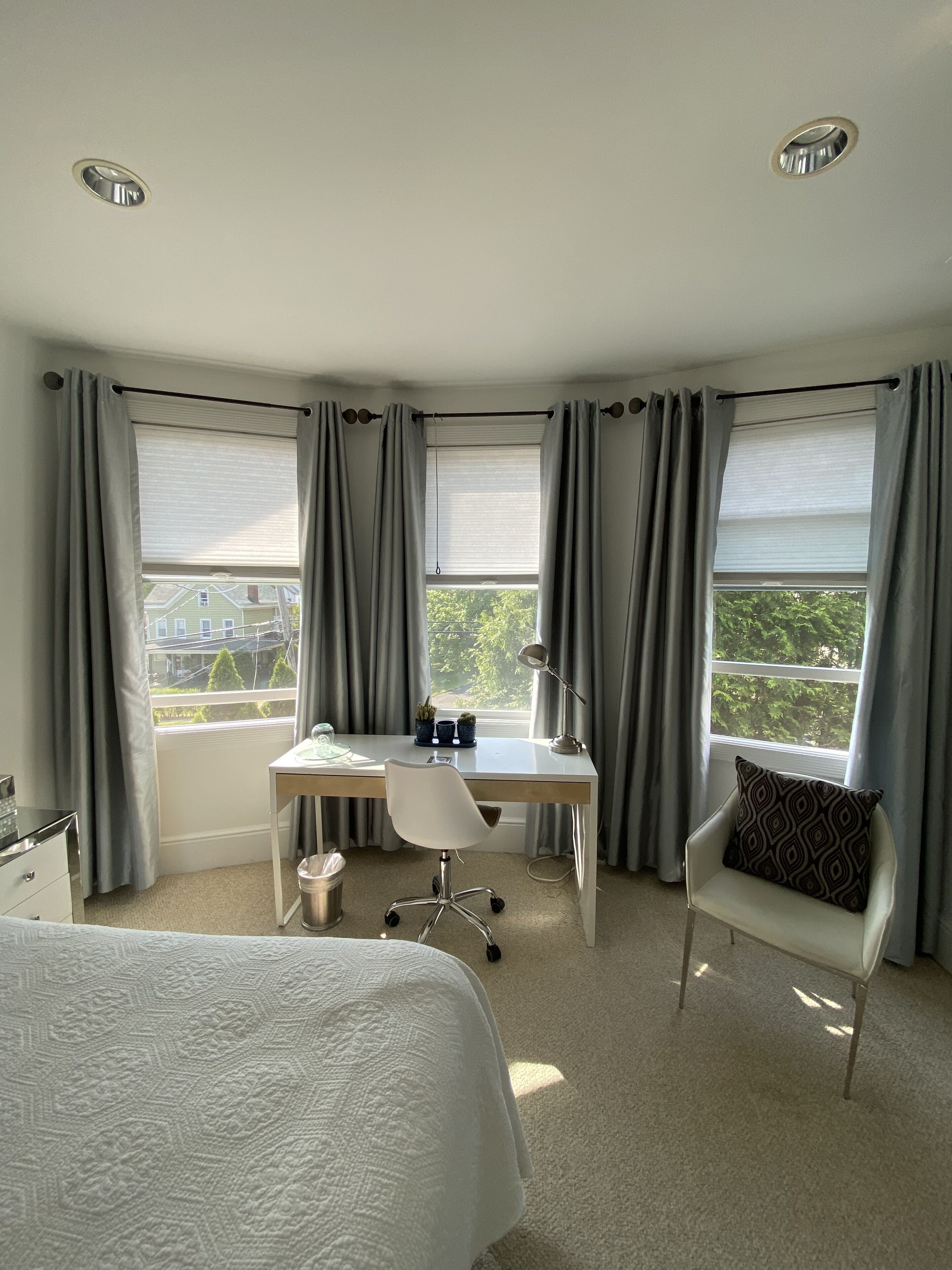 A hotel room with a bed, a desk, a chair, and two windows with curtains.