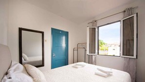 Quarto de hotel com cama, porta azul, janela com cortinas e espelho na parede.