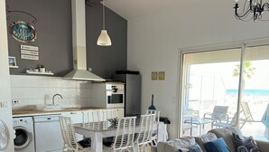 Une cuisine moderne avec des appareils électroménagers blancs, un coin repas avec des chaises blanches et un salon avec un canapé gris et des coussins bleus.