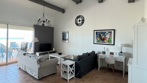 Un salon spacieux avec une grande télévision, un canapé, une table à manger et un bureau. Une fenêtre offre une vue sur la plage.