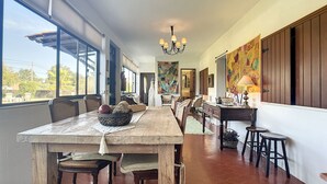 Een eetruimte met een lange houten tafel, stoelen en een kroonluchter. Links is een raam en er is een deur naar een andere kamer.