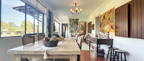A dining area with a long wooden table, chairs, and a chandelier. There is a window on the left side and a door leading to another room.